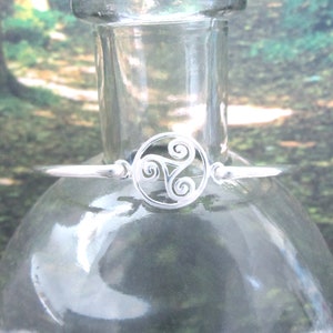 May include: A silver-colored bracelet with a Celtic triskele design. The bracelet features a circular centerpiece with three swirling arms, connected to a thin band. The bracelet is displayed on a clear glass bottle.