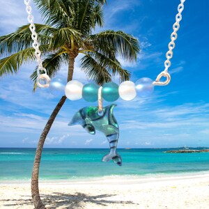 May include: A silver chain necklace with a blue and green dolphin charm and iridescent blue and white beads. The necklace is photographed against a tropical beach scene.