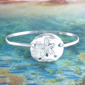 May include: A silver-toned bangle bracelet featuring a sand dollar charm. The sand dollar has a detailed star pattern in the center and several small holes. The bracelet is set against a blurred ocean background.