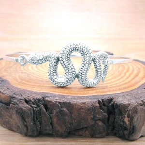 May include: A silver-colored bangle bracelet featuring a detailed snake design. The snake is coiled in a figure-eight shape, with textured scales and a detailed head. The bracelet is displayed on a wooden surface.