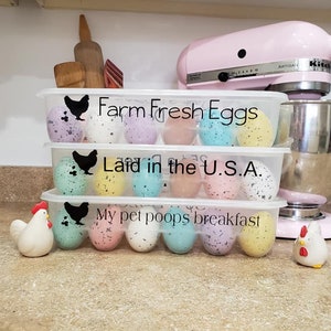 May include: Three clear plastic egg containers with speckled eggs in various colors. The containers have black chicken silhouettes and text that reads "Farm Fresh Eggs Laid in the U.S.A. My pet poops breakfast". Two ceramic chicken figurines and a pink stand mixer are also visible.