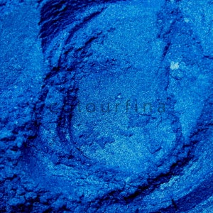 May include: Close-up of a deep blue powder with a sparkly, glittery texture. The powder is a vibrant shade of blue and has a fine, smooth consistency.