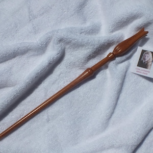 Puede incluir: Una varita de madera marrón lisa y pulida con un mango cónico y una punta acampanada en forma de tulipán. Junto a la varita hay una pequeña tarjeta rectangular con un retrato y el texto impreso LUNA LOVEGOOD MAGIC WAND.