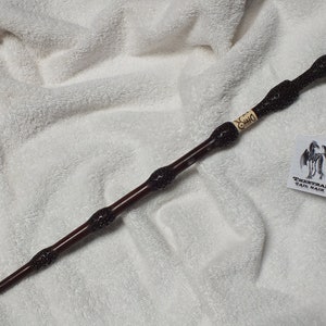 May include: A dark brown wooden wand with black accents and a silver tip. The wand is decorated with a black and white design. The wand is labeled "Draco".