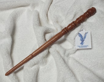 May include: A wooden wand with a brown finish and a rounded tip. The wand has a series of raised bumps along its length. The wand is lying on a white, fluffy surface. A tag with the text "Ice Phoenix Feather" is visible in the background.