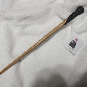 Puede incluir: Una varita de madera con un mango oscuro y un eje marrón claro. Una pequeña tarjeta con la imagen de una persona y el texto "Ron Weasley Wand" está junto a la varita. La varita está sobre una superficie blanca texturizada.
