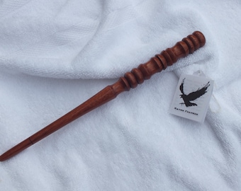 May include: A handcrafted wooden wand with a dark brown finish. The wand features a detailed handle with carved rings and a tapered tip. A small tag with a raven graphic and the text "Raven Feathers" is attached.