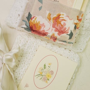 May include: A white crocheted blanket with a floral print fabric pouch and a card with the text "I love those who love me, and those..." and a card with a floral illustration and the text "A generous person will prosper: whoever refreshes others will be refreshed. Proverbs 11:25"