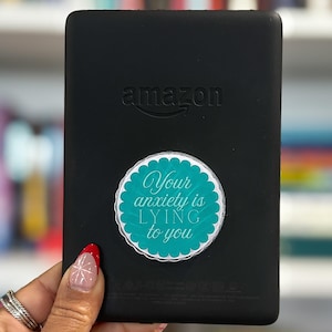 Puede incluir: Un lector de libros electrónicos Kindle negro de Amazon, con el logotipo de la marca. Una pegatina redonda con el texto "Your anxiety is lying to you" está adherida. El lector se sostiene en una mano con un diseño de uñas rojo y blanco.