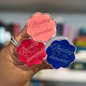 May include: Three colorful, flower-shaped acrylic pins. Each pin has the phrase "Reading is Fun-damental" in a script font. The pins are in shades of pink, red, and blue. The background is a blurred image of a bookshelf.