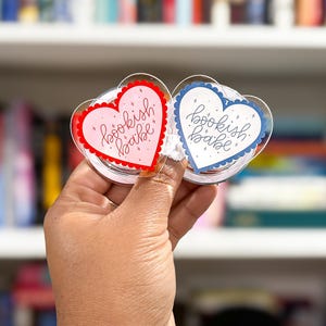May include: Two heart-shaped book accessories with the text "bookish babe." One is pink with a red border, and the other is blue with a blue border. The accessories are held up in front of a bookshelf.