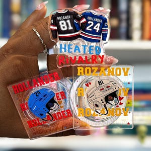 May include: Clear acrylic hockey-themed accessories featuring jersey designs with player names and numbers, and helmet designs. The text "HEATED RIVALRY" and player names are visible. The jerseys are black and blue.