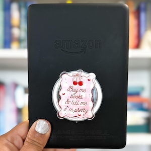 May include: A black Amazon Kindle e-reader with a pink and white pop-up phone grip. The grip has a cherry design and the text "Buy me books & tell me I'm pretty". The e-reader is held in a hand.