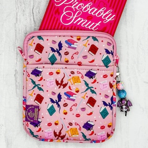 Puede incluir: Bolso bandolera rosa con un estampado de fantasía de dragones, libros, espadas y labios. Dentro hay un cuaderno rosa con el texto "Probably Smut". Un charm con cuentas y un dragón está adjunto.