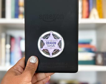 Könnte beinhalten: Ein schwarzer Amazon E-Reader mit einem kreisförmigen, hellvioletten, sternförmigen Zubehörteil auf der Rückseite. Das Abzeichen zeigt den Text "DRAGON tamer" in dunkelvioletten Buchstaben, umgeben von einem weißen und violetten Rand.