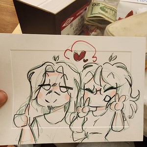 Peut inclure: Un croquis de deux personnages de dessins anim&eacute;s, l'un faisant un pouce lev&eacute; et l'autre un signe de paix. Le dessin est &agrave; l'encre noire et verte avec des accents rouges, dans un passe-partout blanc.