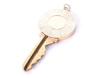 May include: A gold-colored key pendant with a circular top and a key-shaped bottom. The circular top has a textured design around a smooth, central circle. The key's teeth are visible, and a small loop is at the top for a chain.