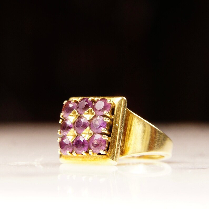 May include: A gold ring with a square-shaped setting featuring a cluster of purple gemstones.