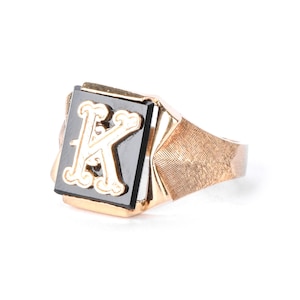 Op de afbeelding: Een goudkleurige ring met een zwarte onyx steen met een verhoogde, sierlijke letter "K" in een lichtere goudkleur. De ring heeft een getextureerde band en een rechthoekige voorkant.