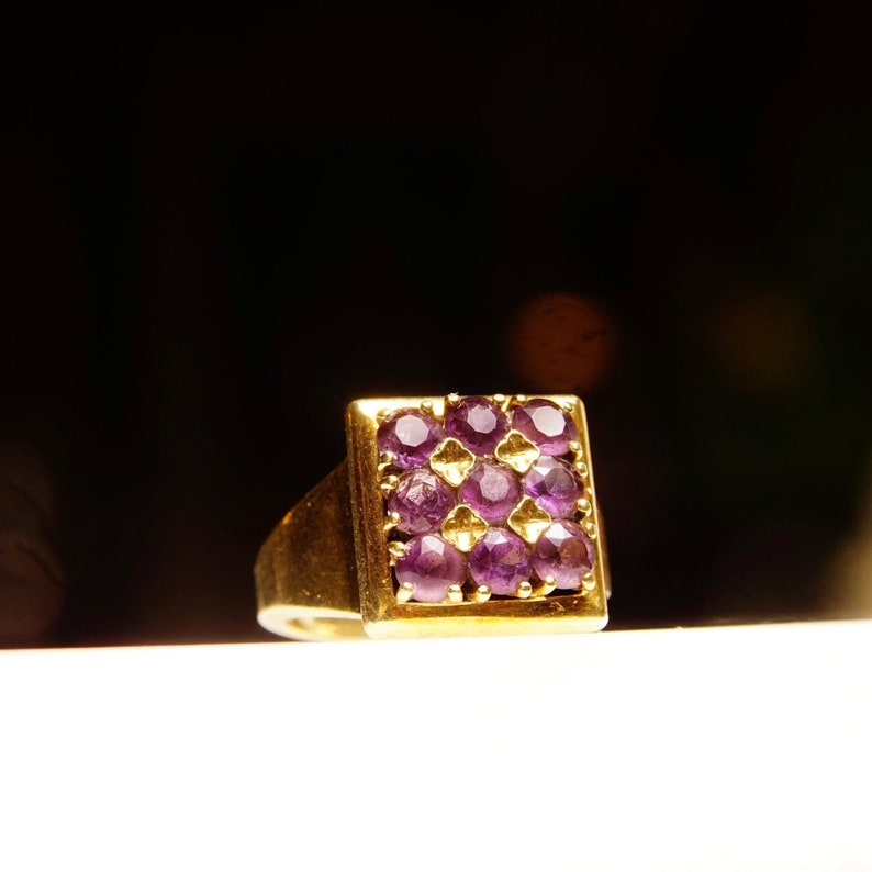 May include: A gold ring with a square-shaped setting featuring a cluster of purple gemstones.
