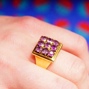 May include: A gold ring with a square-shaped setting featuring a cluster of purple gemstones.