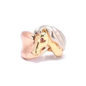 Puede incluir: Un anillo de oro con un diseño de cabeza de caballo. La cabeza del caballo es dorada con un ojo de gema roja y una melena plateada. La banda del anillo es de color oro rosa.