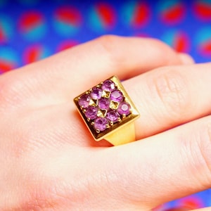 May include: A gold ring with a square-shaped setting featuring a cluster of small, purple gemstones.
