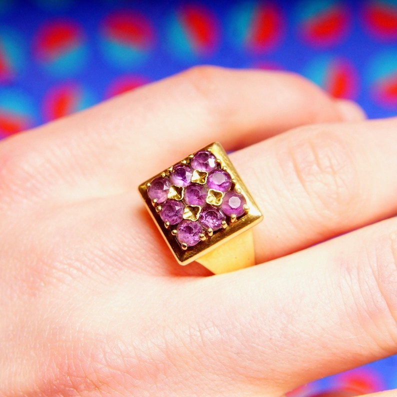 May include: A gold ring with a square-shaped setting featuring a cluster of small, purple gemstones.