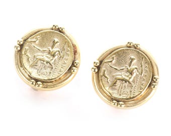 May include: A pair of gold-colored stud earrings. Each earring features a circular design with a detailed relief of a seated figure holding an object, surrounded by text. Small gold beads adorn the outer edge.