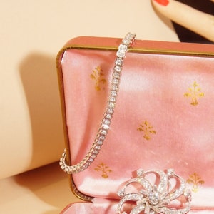 May include: A delicate diamond tennis bracelet, featuring a continuous line of sparkling round diamonds set in a white metal band. The bracelet is displayed in a pink velvet jewelry box.