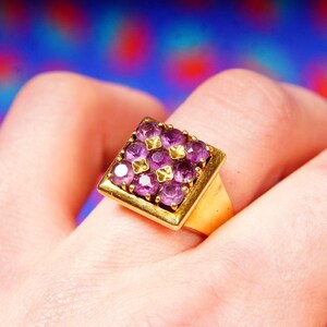 May include: A gold ring with a square-shaped setting featuring a cluster of purple gemstones.