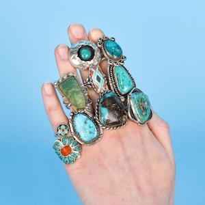 May include: A hand showcasing a selection of silver rings, each set with distinctive gemstones. The rings incorporate turquoise, green, and other vibrant stones in diverse forms and mountings. The backdrop is a plain light blue.
