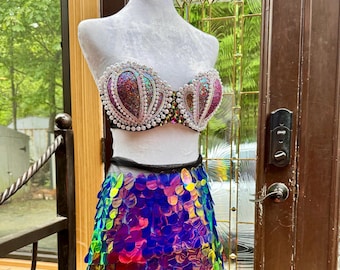May include: A mermaid-themed costume displayed on a mannequin. The top is a bedazzled bra with iridescent sequins and pearl accents. The skirt is made of iridescent, colorful, scale-like sequins. The costume is designed for a theatrical or fantasy setting.