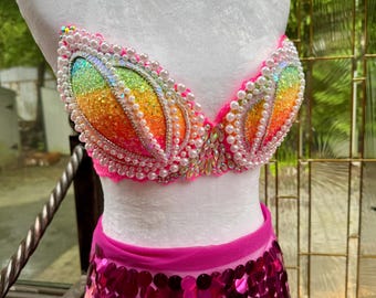 May include: A vibrant, handmade bra and skirt set. The bra features a rainbow-colored design with iridescent sequins and pearl accents. The skirt is covered in pink sequins. The set is displayed on a white mannequin.