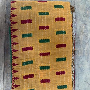 May include: Folded mustard-colored quilt with a repeating pattern of green and burgundy rectangles, stitched with white thread. The quilt's edge features a decorative border. A cozy, handmade textile.