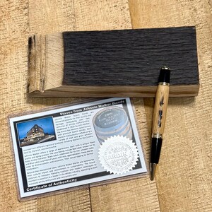 May include: A wooden pen with a black tip and accents, lying on a wooden block. A certificate of authenticity is next to the pen, with text that reads "Staves from Johnnie Walker Blends". The wood has a dark, charred appearance.