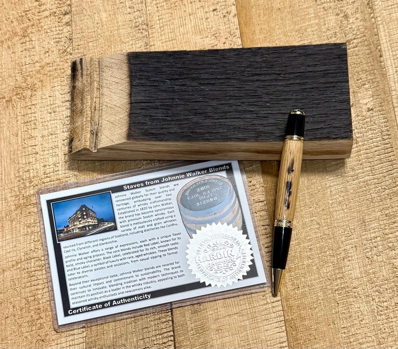 May include: A wooden pen with a black tip and accents, lying on a wooden block. A certificate of authenticity is next to the pen, with text that reads "Staves from Johnnie Walker Blends". The wood has a dark, charred appearance.
