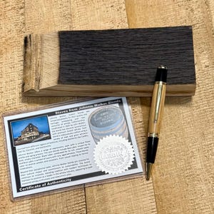 May include: A wooden pen with gold accents and black tips rests on a dark wood block. A certificate of authenticity is visible, with the text "Staves from Johnnie Walker Blends" and a picture of a building.