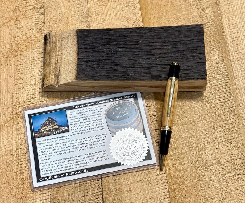 May include: A wooden pen with gold accents and black tips rests on a dark wood block. A certificate of authenticity is visible, with the text "Staves from Johnnie Walker Blends" and a picture of a building.