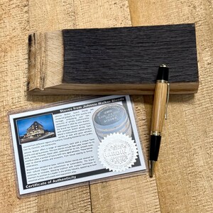 May include: A wooden pen with gold accents rests on a wooden surface next to a piece of charred wood and a certificate. The certificate reads "Staves from Johnnie Walker Blends" and includes a photo of a building.