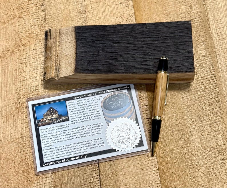 May include: A wooden pen with gold accents rests on a wooden surface next to a piece of charred wood and a certificate. The certificate reads "Staves from Johnnie Walker Blends" and includes a photo of a building.