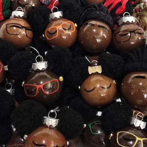 May include: A collection of round, brown, glossy Christmas ornaments featuring black pom-pom hair. Each ornament displays painted facial features, including closed eyes with long lashes and various colored glasses or a small gold glitter bow accessory.