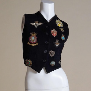 Puede incluir: Un chaleco de terciopelo negro con una variedad de parches bordados de colores. El chaleco tiene cierre de botones y una longitud recortada.