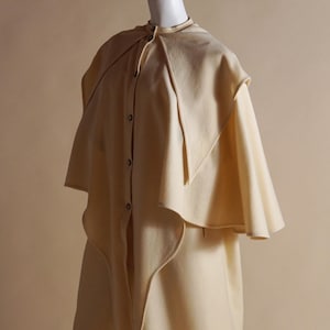 May include: A cream-colored wool cape with a button-up front and a wide, flowing collar. The cape has a unique design with overlapping layers of fabric.