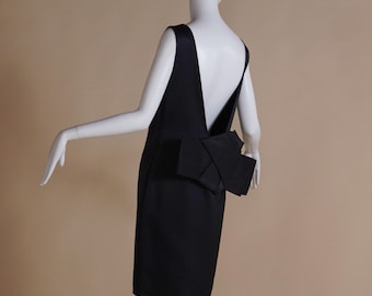 May include: A black, sleeveless dress, reaching the knees, with a low V-shaped back and a prominent bow feature at the lower back. The dress is presented on a mannequin against a plain backdrop, highlighting its understated elegance.