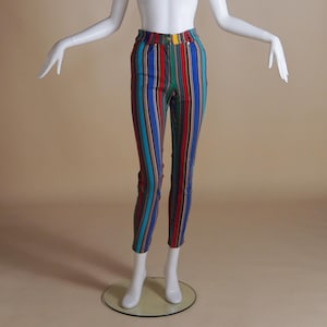 May include: A mannequin wearing striped trousers. The trousers have vertical stripes in red, blue, green, and teal. They have a high waist and a yellow button. The mannequin is white and faceless. The trousers' measurements are in centimetres.