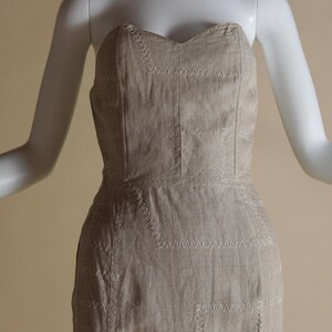 May include: A strapless dress made of a light brown linen fabric with white stitching. The dress has a fitted bodice and a straight skirt.