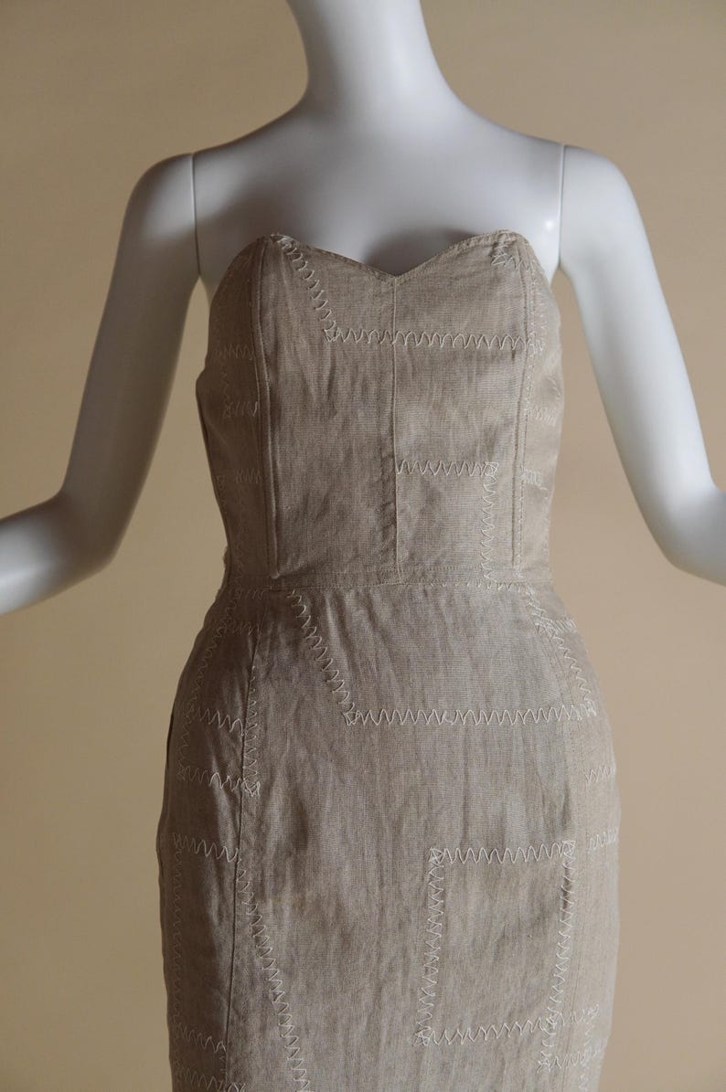 May include: A strapless dress made of a light brown linen fabric with white stitching. The dress has a fitted bodice and a straight skirt.