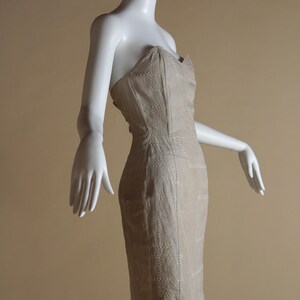 May include: A strapless dress in a light beige colour with a geometric pattern of white stitching. The dress is fitted and has a short length.
