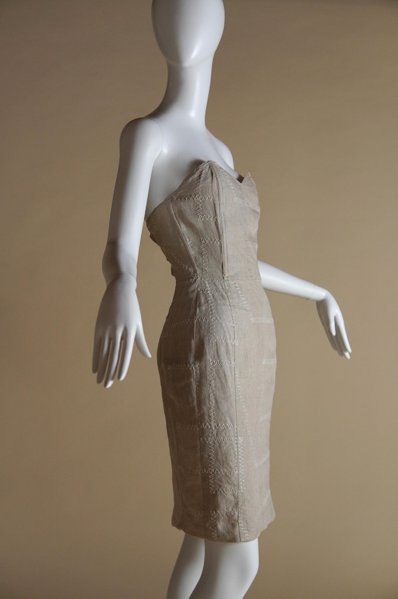 May include: A strapless dress in a light beige colour with a geometric pattern of white stitching. The dress is fitted and has a short length.